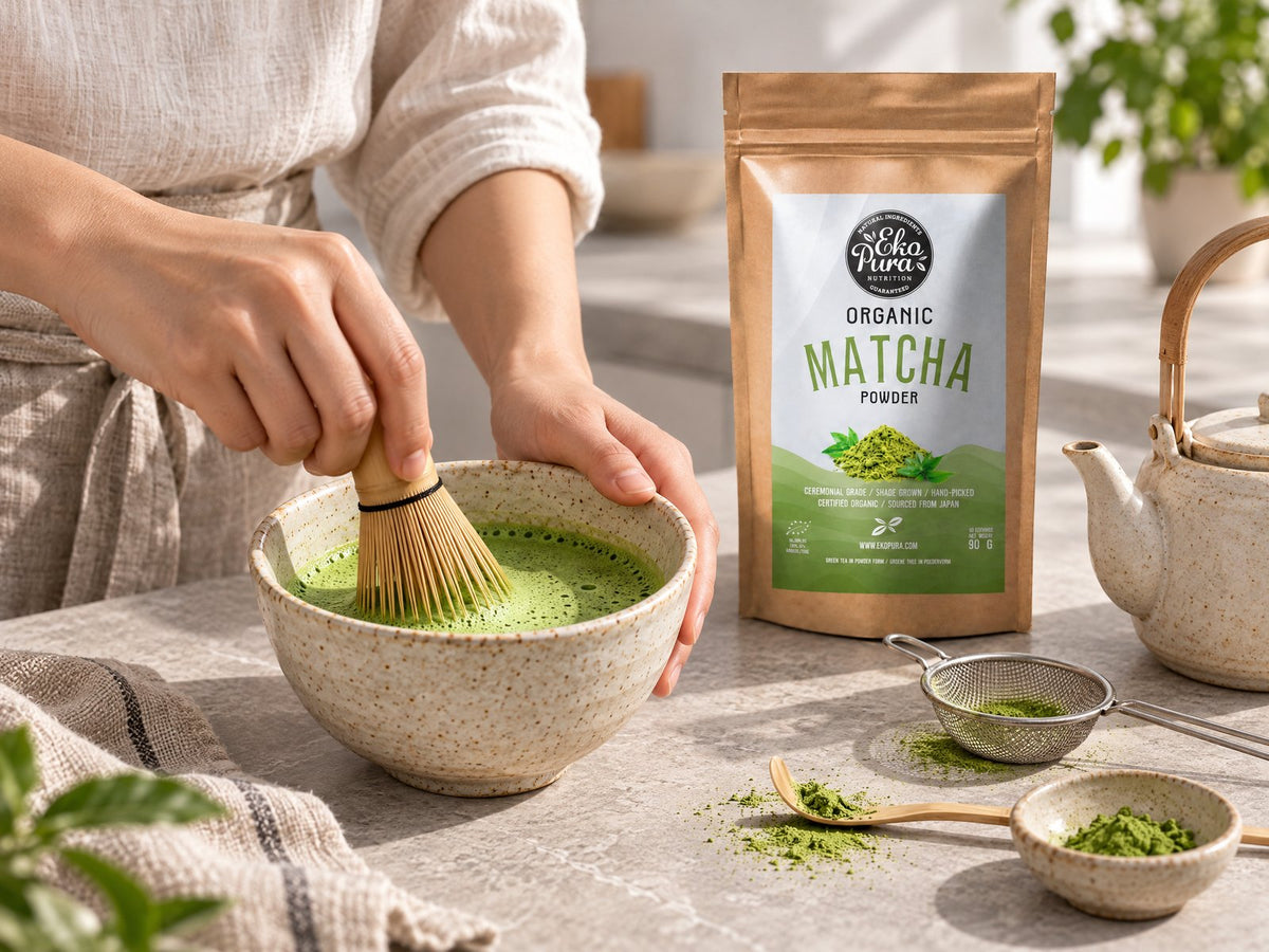 Matcha wordt met een bamboe whisk opgeklopt in een kom, naast een zeefje, een bamboe lepeltje en Ekopura Matcha Poeder Bio